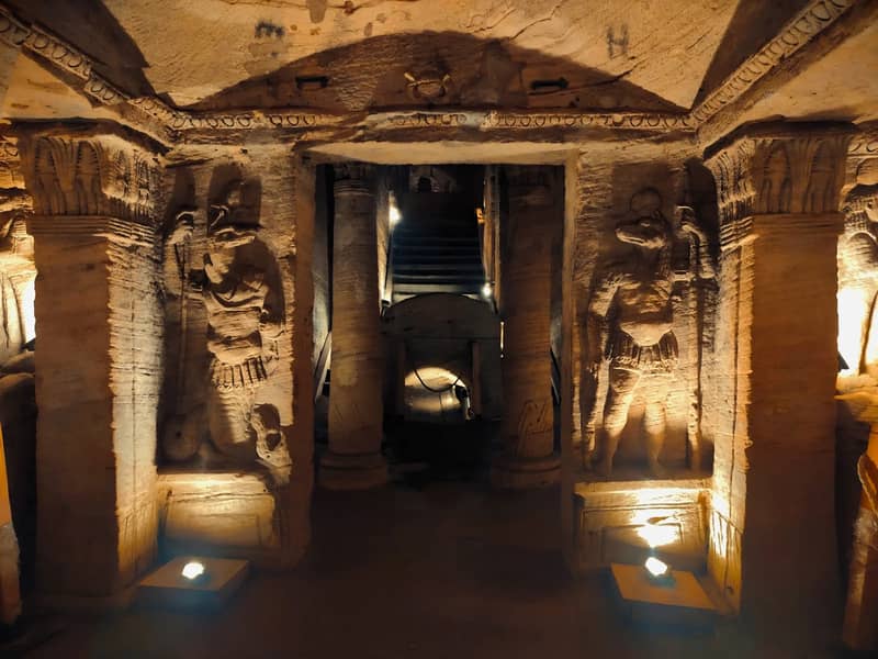 Cámara funeraria excavada en la roca e iluminada, con figuras talladas y columnas, en las catacumbas de Kom el Shoqafa